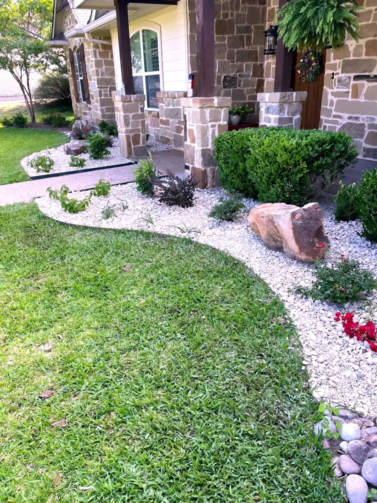 A stone home in Central Texas with white rock beds, shaped shrubs, a boulder accent, and flowering color installed by H&M.