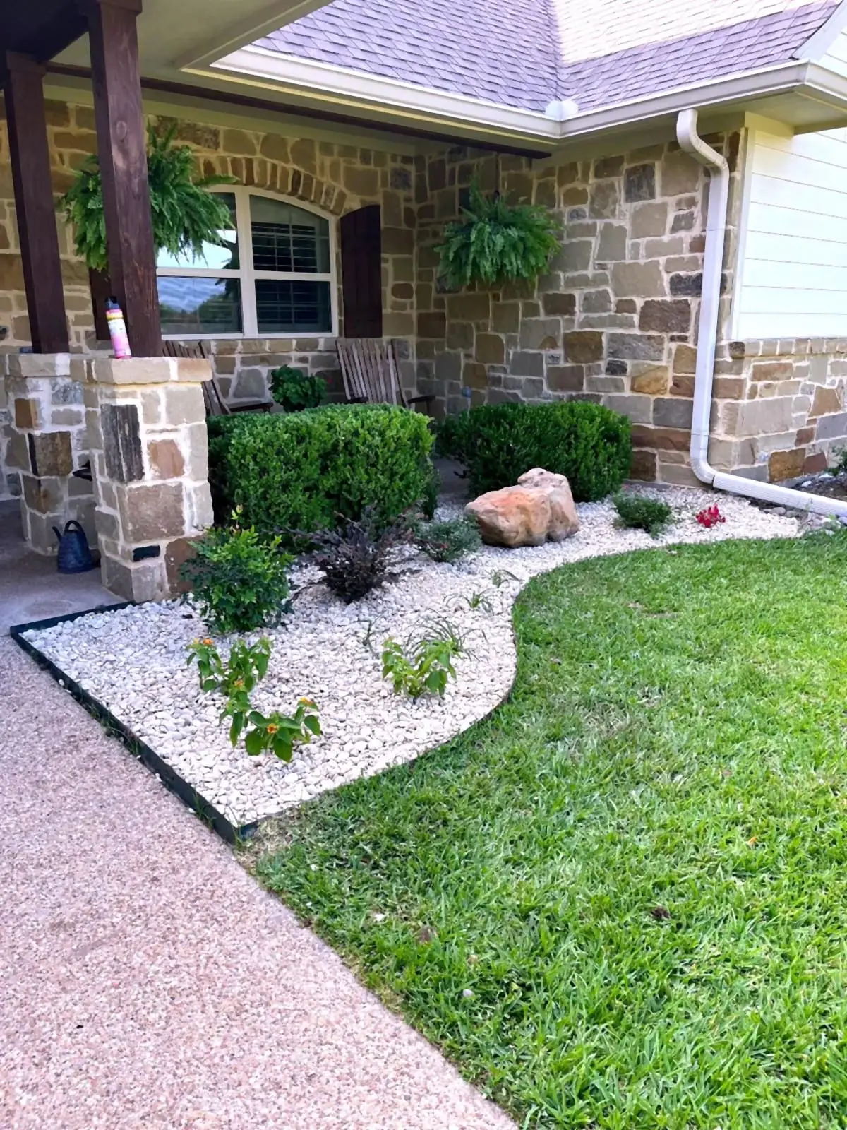 A stone home with white river rock beds, boulder accents, and clipped shrubs beside turf.